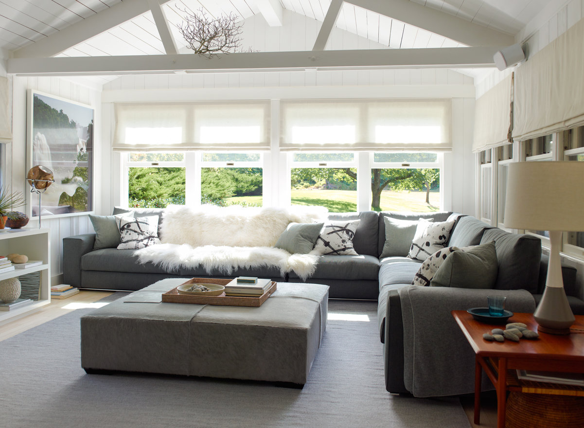 Den in white and gray palette with white wood ceiling, windows with view of green lawn and trees, gray sofas and ottoman.