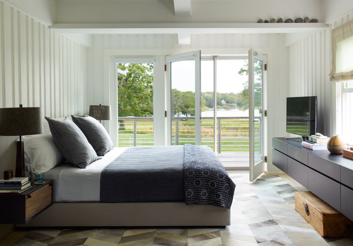 Bedroom with chevron hide rug, bed with gray cover, wall-mount dresser, glass door open terrace and with lawn and water view.