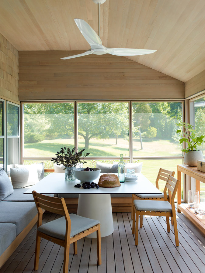 Porch with wood ceiling and white fan, wood plank floor, banquette seat with gray cushion pedestal table and wood chairs.