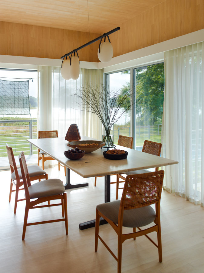 Dining room with pale wood ceiling and floor, wood table and chairs, hanging light, sliding glass doors with sheer curtains.