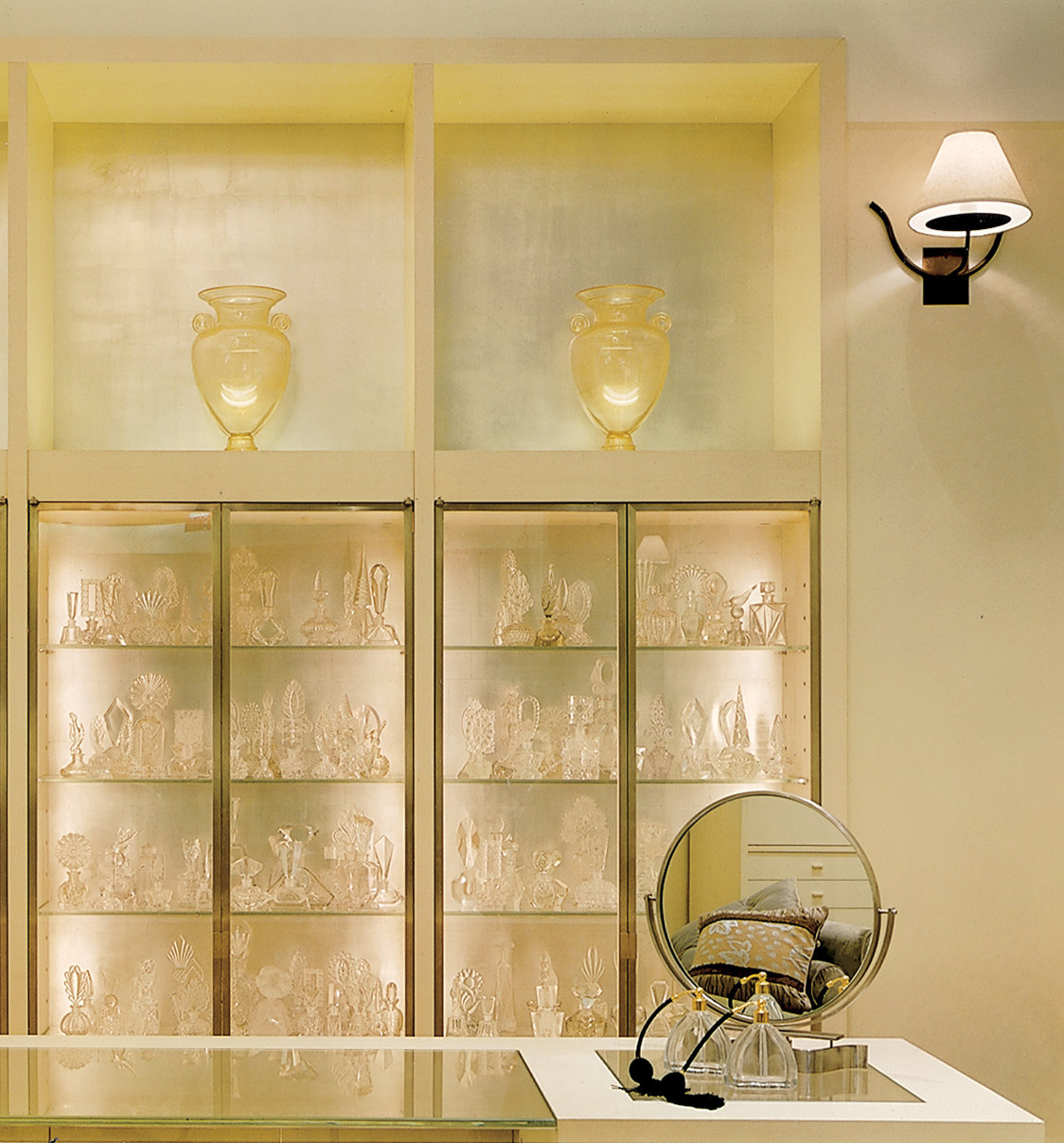 Built-in wall display in a high-end store with platinum-colored walls, and glass front shelves that hold decorative glass perfume bottles.