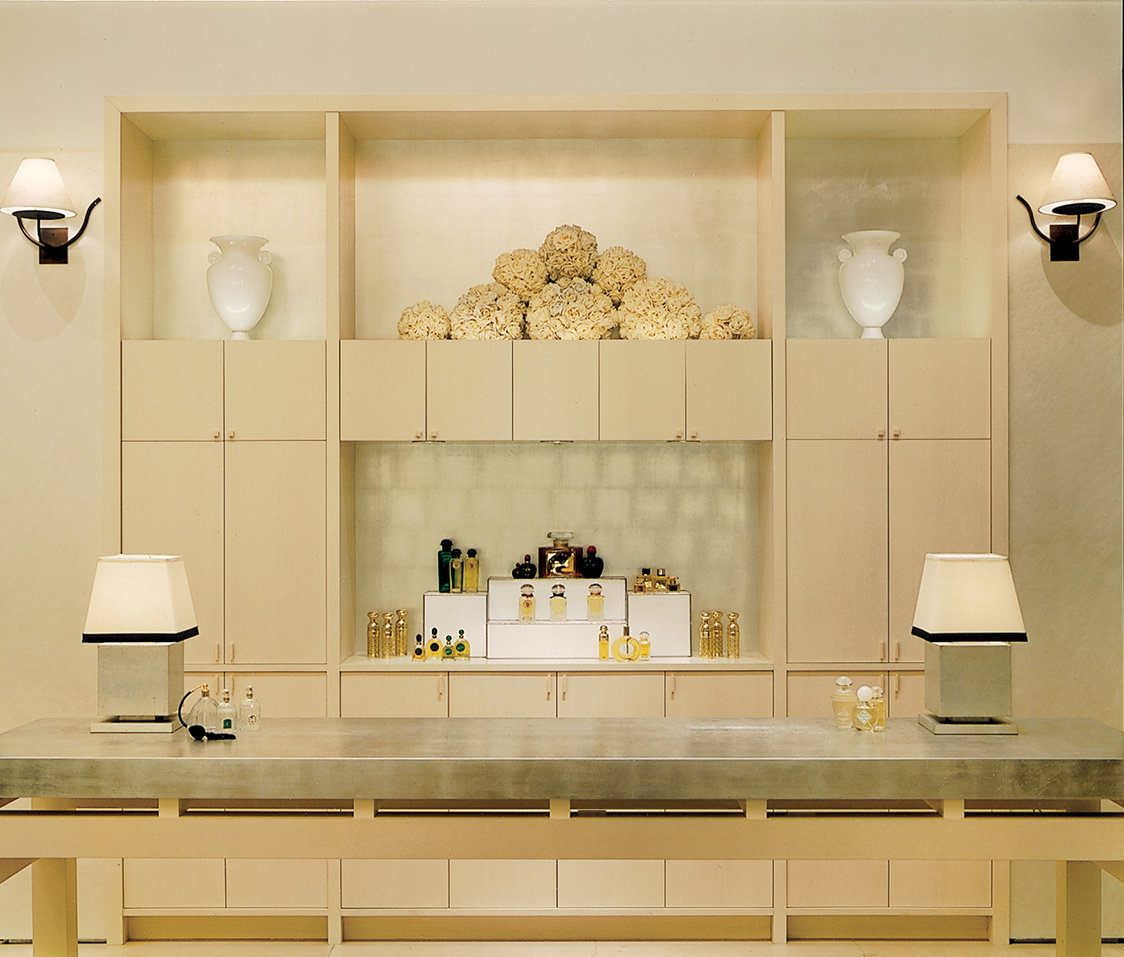 Display wall of a high-end store in a neutral palette with platinum-colored walls, blonde wood cabinetry and shelving with perfume bottles.