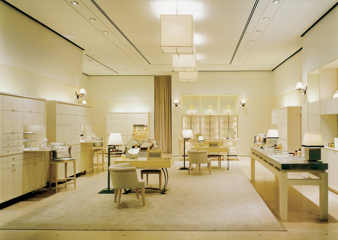 Light neutral store interior with blonde wood cabinetry, shelving and tables, beige marble floor and beige carpet and light fixtures with white shades.