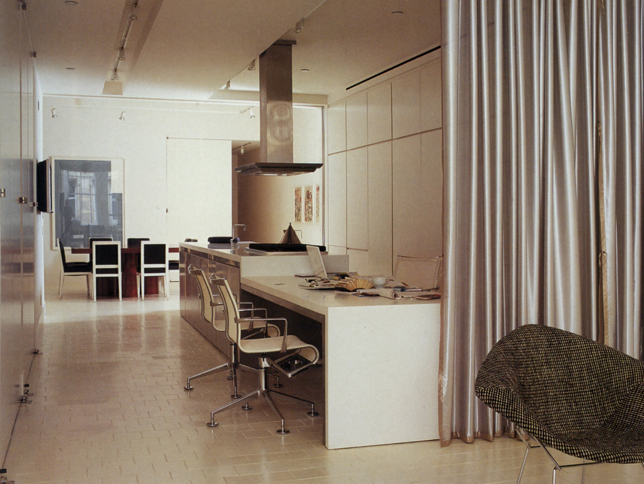 Open kitchen-office in white with modern white desk; dining table and black chairs in background, silver curtain in foreground.
