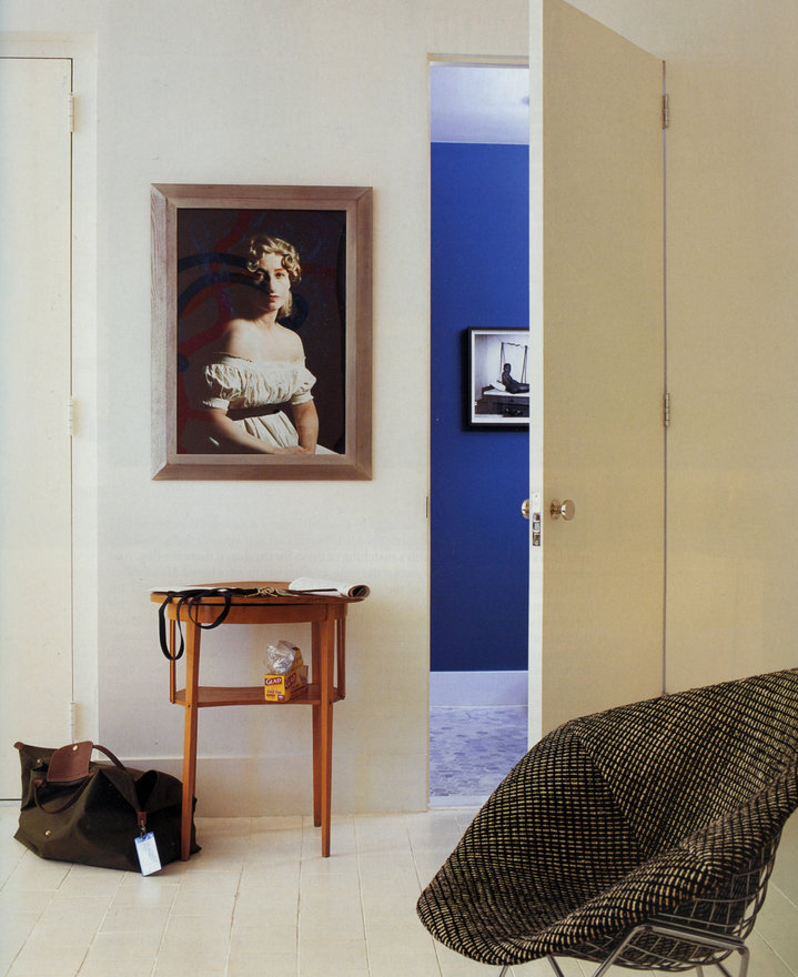 White room with framed portrait of a woman on the wall, small wooden table, tote bag on floor and door open to a blue room.