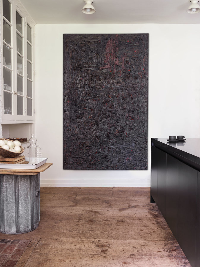 Large black painting on white wall of kitchen, white cabinet with glass front to left, black counter to right, wood floors.