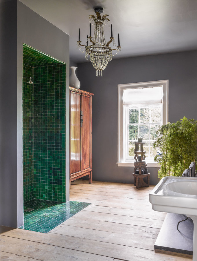 Bathroom with gray walls, wood plank floor, green tile standing shower, crystal chandelier, window and white pedestal sink.