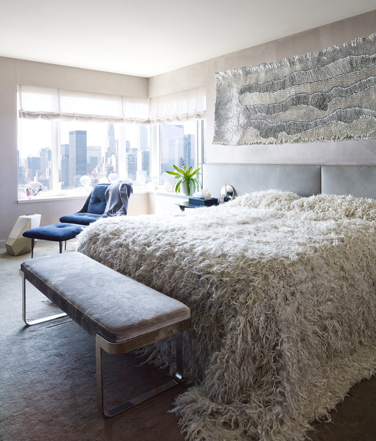 Airy bedroom with silver textile wallhanging, white Mongolian fur bedspread, grey velvet bench, and blue velvet lounge chair.