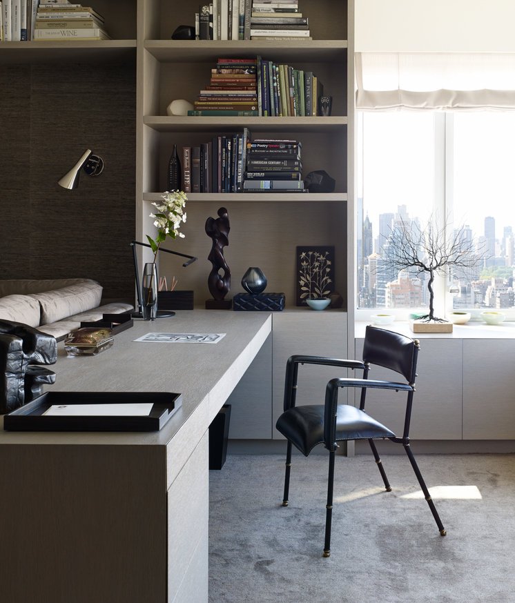 Home office with pale wood shelving, cabinetry and desk, light grey carpet, black leather armchair, window in background.