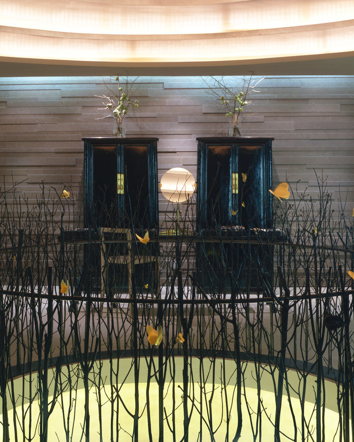 Close-up of a circular railing constructed of bronze twigs that surrounds an opening in the floor. Back wall with a pair of black Chinese armoires.