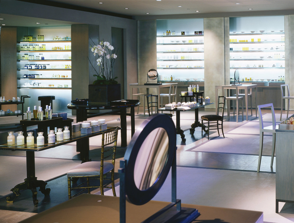 Interior of a cosmetics and fragrances store with gray walls and flooring, backlit display shelves and black lacquer furniture.