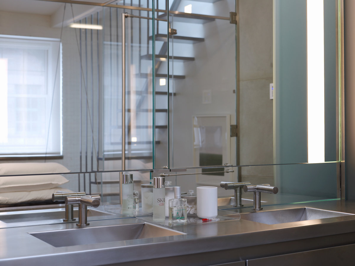 Close-up inside bathroom with metal double sink vanity with modern faucets, mirror showing reflection of window and staircase.