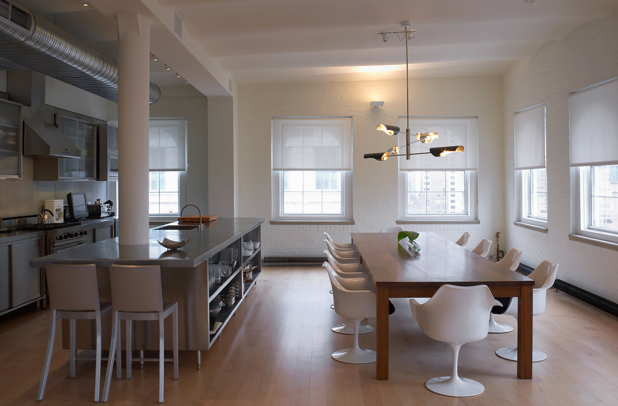 Modern, open stainless-steel kitchen with island and stools, dining room with wood table and white chairs, two window walls.