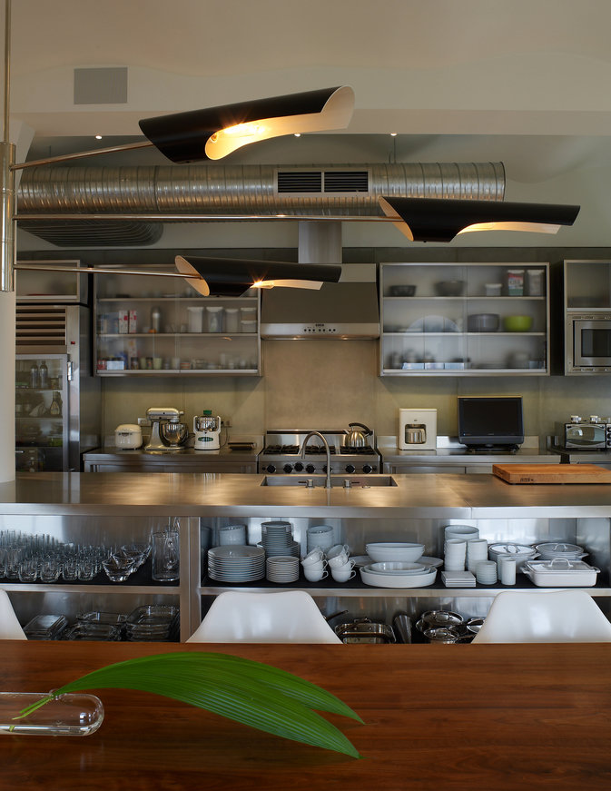 Modern kitchen with stainless steel counter and island with shelving for glassware and dinnerware; wood table in foreground.