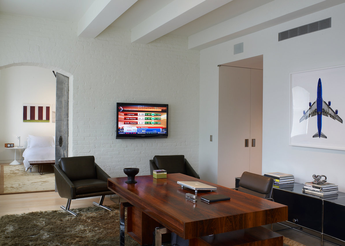 Home office with wood desk, black leather chairs, area rug, white brick walls with arched doorway to bedroom, wall mount tv.