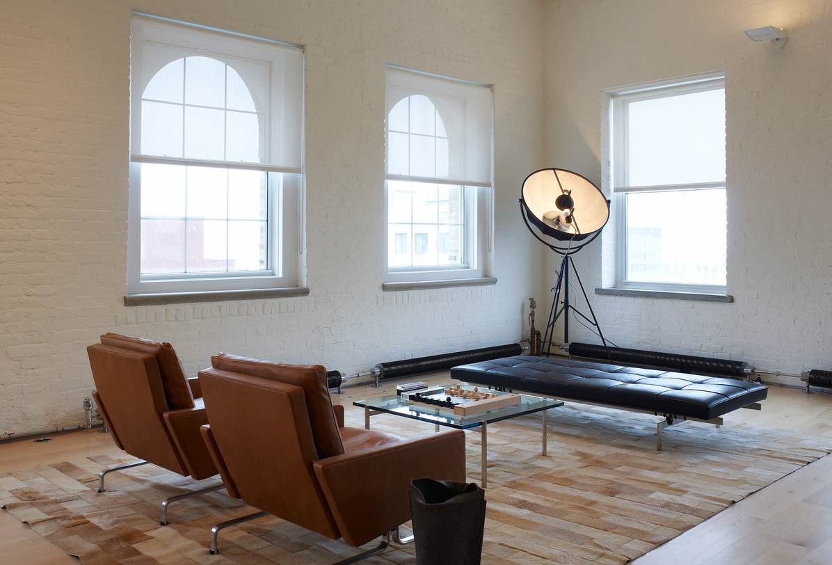 Living room with white brick walls, wood floor, white windows, tripod light, leather chairs and bench and glass coffee table.