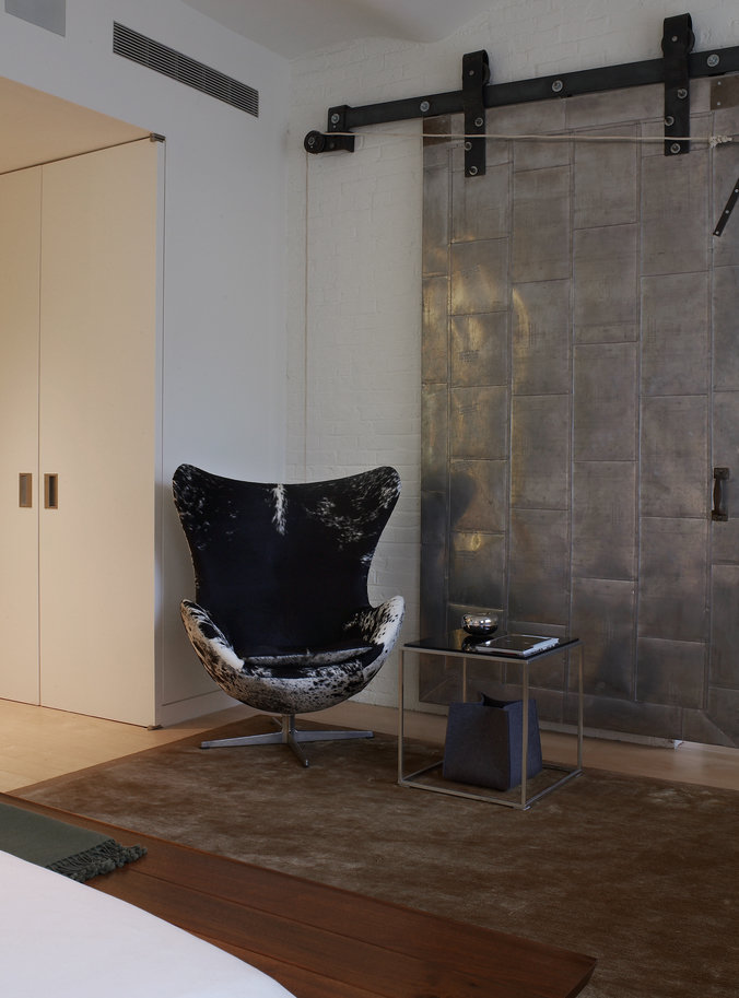 Bedroom with egg chair upholstered in black and white hide, metal barn door, tan area rug, bed and wood bench in foreground.