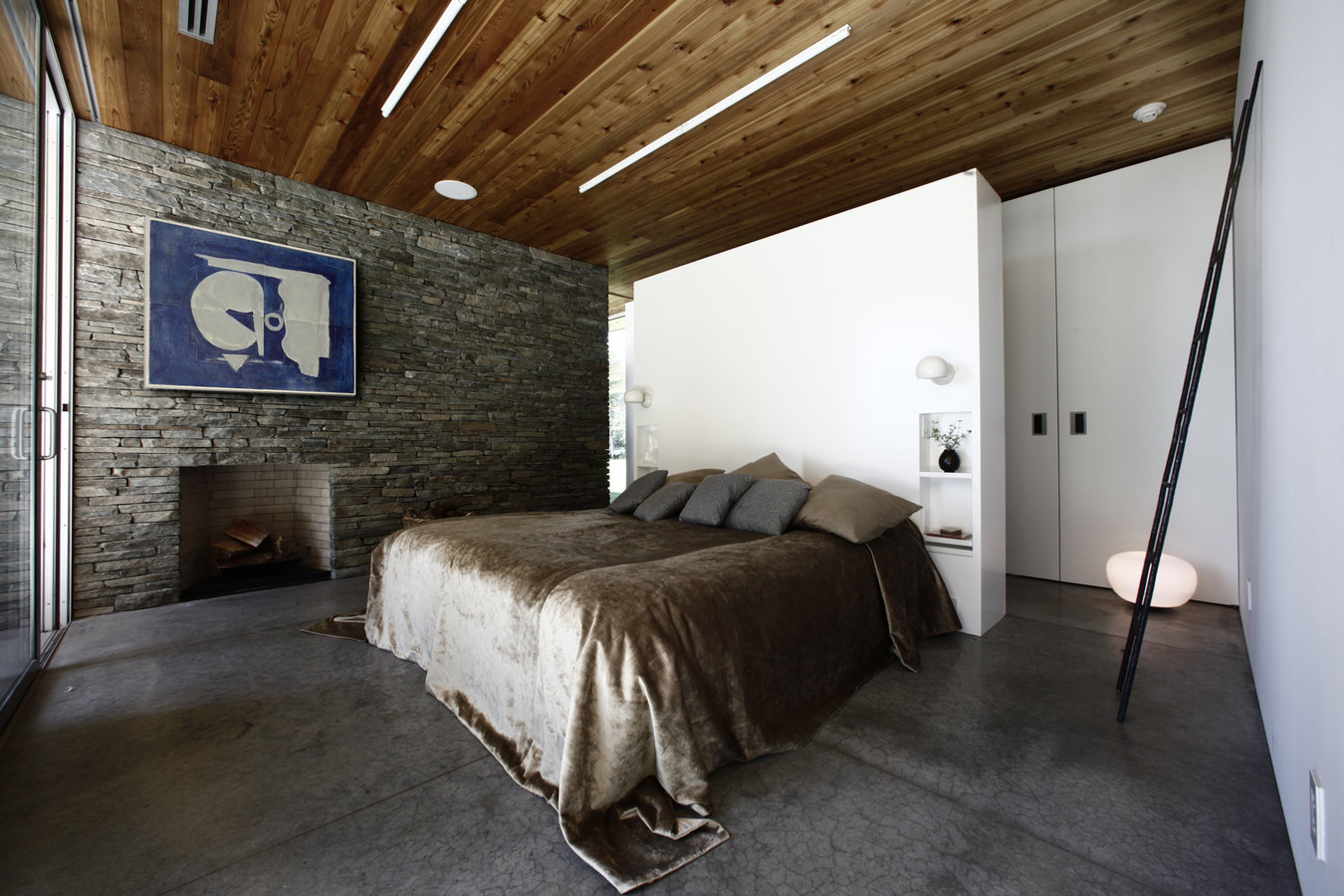 Bedroom with stone wall, fireplace, blue and white art, cedar ceiling, white walls, concrete floor and bed with velvet cover.