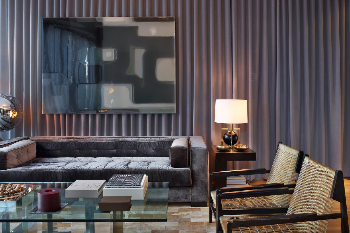 Living room with gray velvet sofa, glass coffee table, woven rope and wood chairs in front of a wall of curtains with art.