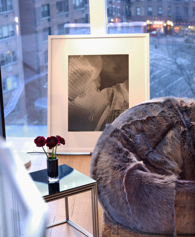 Fur beanbag, small square open frame side table with flowers in vase, framed photograph on floor leaning against windows.